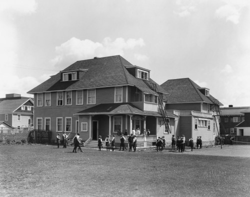 Black and white image of children in school uniform playing outside in front of school building