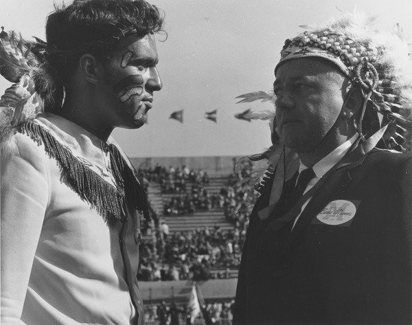 Robert Bertram in Native American headdress, ca. 1980. Robert E. "Bob" Bertram of the Class of 1949 wears a Native American headdress while standing with an unidentified man, also in Indian dress, at McGuirk Stadium. Photo titled "Metawampi".