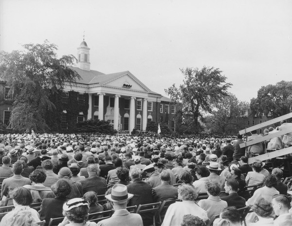 Commencement at Goodell Library, ca. 1955. A crowd of spectators, some in seated in bleachers, listen to a speaker as Commencement ceremonies are held in front of Goodell Library (possibly in 1955).