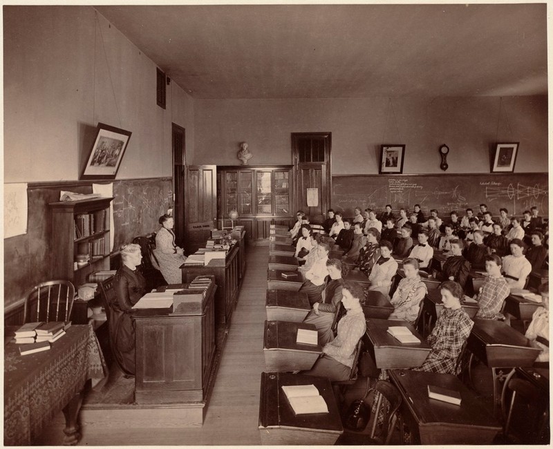 Black & White photo of young women in long-sleeved, long dresses at rows of individual desks as teachers lecture from the front of the classroom.