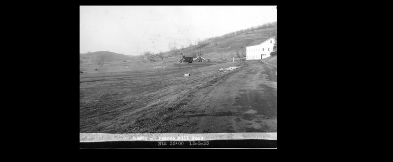 Black and white image showing a dirt road with a large white barn on the right side, with a field and construction equipment on the left side in the distance.