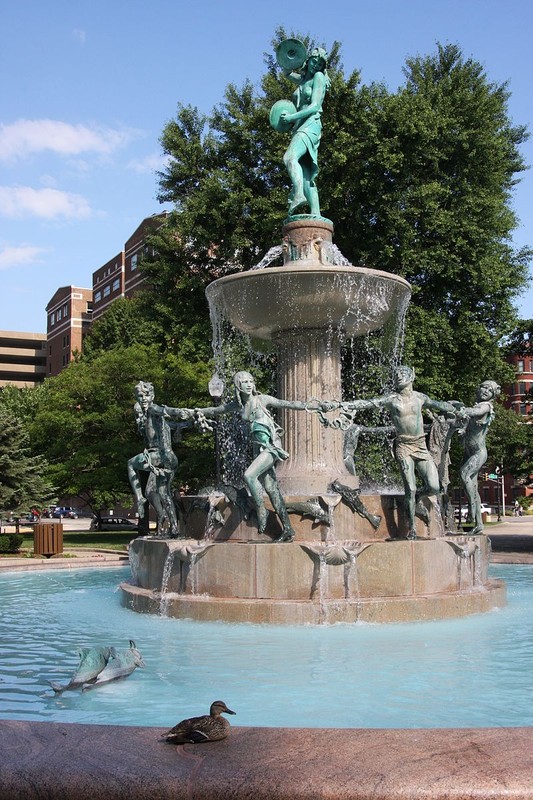 This bronze sculpture features eight children around the base and is topped by a woman who is playing cymbals and dancing with the children below. 