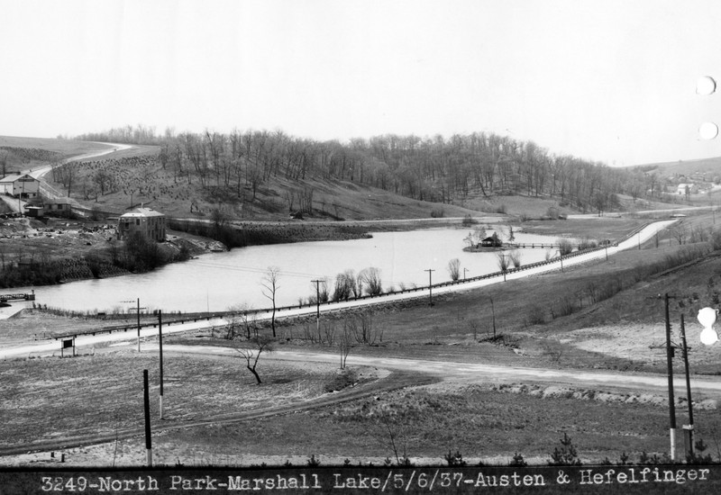 Black and white photo of a lake, taken from a nearby hill. There is a small island at the center, connected to the shore by a small footbridge. On the distant shore is a stone building.