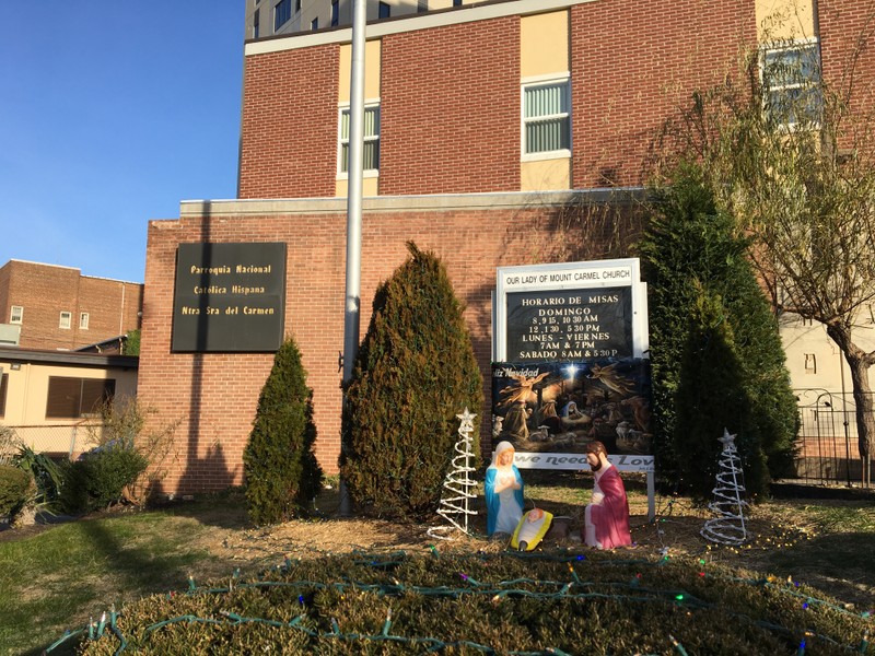 Front lawn view, more Christmas lights on trees and bushes, manger set up, and poster with the words "Feliz Navidad"