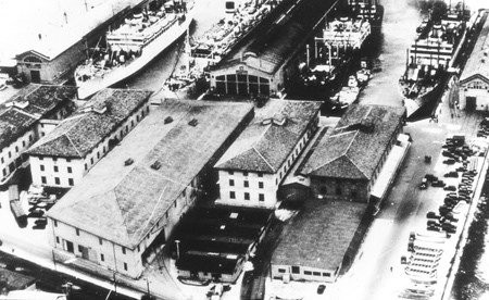 This photo shows the San Francisco Port of Embarkation in action: 4 large army transport ships are docked alongside the loading piers, ready to accept goods from the adjacent warehouses. Photo circa 1920s.