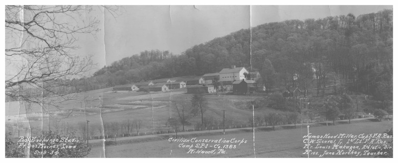 Black and white landscape photo, taken across Wildwood Road, showing a group of buildings on the hillside. It includes eight long one-story barracks, a two level barn, three houses, and an additional one-story structure.