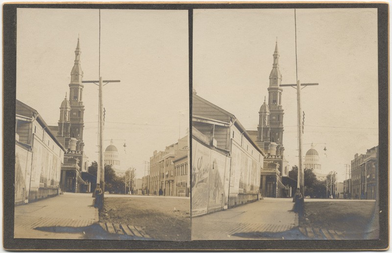 The unpaved streets of late 19th Century Sacramento can be seen in this stereograph from 11th and K Streets, from which the spire of the Cathedral can be seen in the background (CA State Library).
