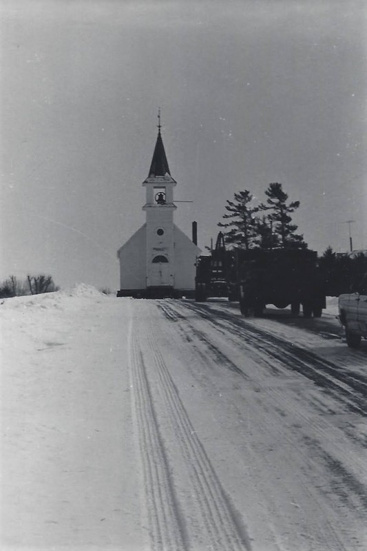 Caravan of vehicles leading the machinery carrying the church along a snowy road.