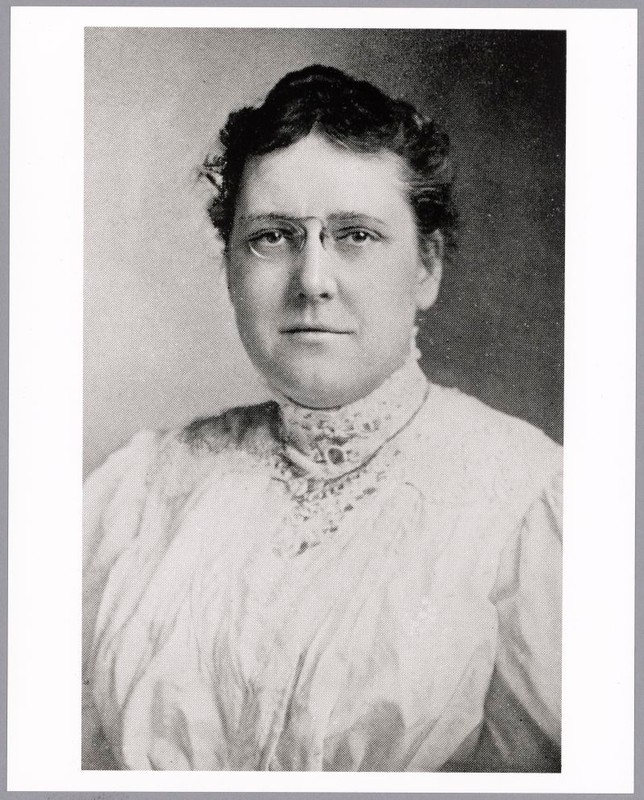 Black & white, woman, dark hair pulled back, light metal rimmed glasses, white blouse with a high lace collar