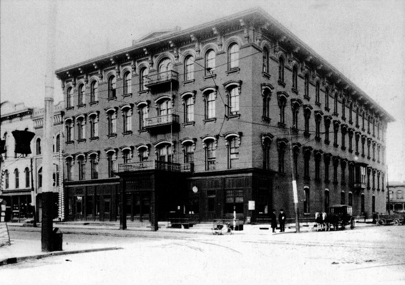 A black and white photo of a street scene. A row of buildings is seen, with carriages and a . Second from left is the C.H. Little Building. Our view of it is partially obscured by a telephone pole, but the plate tower is clearly visible. At right is the Brewster House, and on the corner in front of it are an early bicycle leaning against a rail, and two men in dark coats. Nearby are a horse-drawn carriage and a parked wagon.