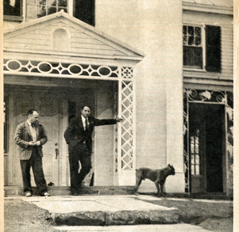Black and white photo of two men standing on the front porch of a house with a dog to the right running away.