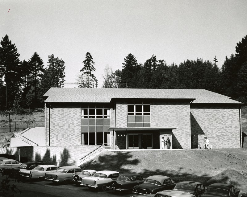 Black and white photograph of the exterior of a midcentury, two-story building, sitting in a canyon with a parking lot filled with 1950s style cars parked in front. Two figures are visible standing at the entrance.