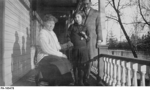 Black and white image of older woman sitting, young girl and man standing, on porch