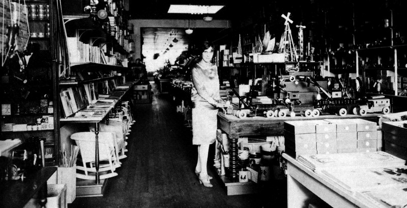 A black and white photo of a store interior circa 1925. The room is narrow and deep and contains many toys and books on shelves and table displays. A young woman stands at center in a blouse and skirt cut just below the knee.