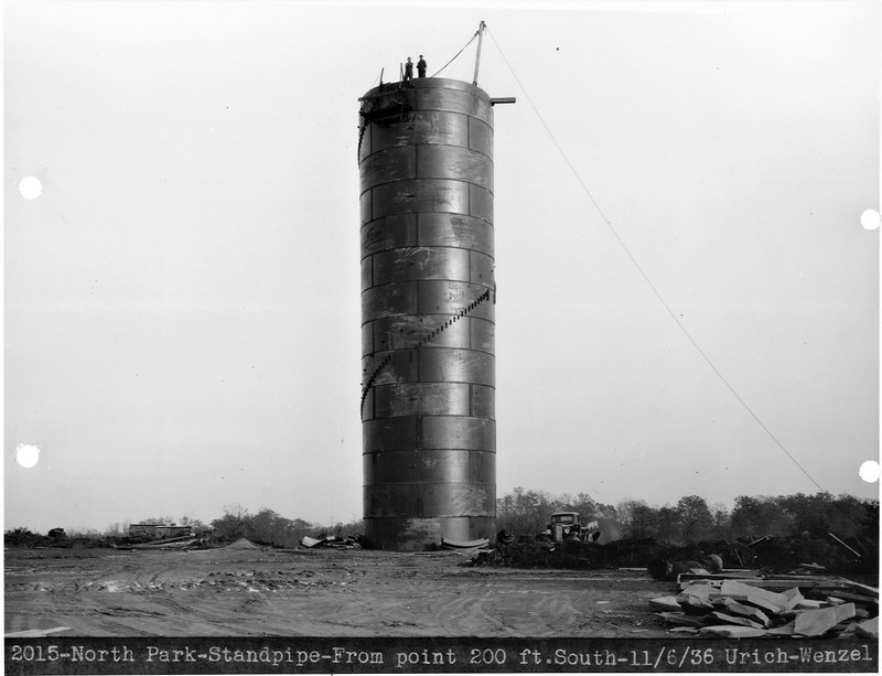 Black and white image shows a large round metal structure, constructed from 14 rows of large metal panels. Each panel is slightly shorter than the pick-up truck parked at its base. Two people are standing at the very top, looking down towards the photographer.