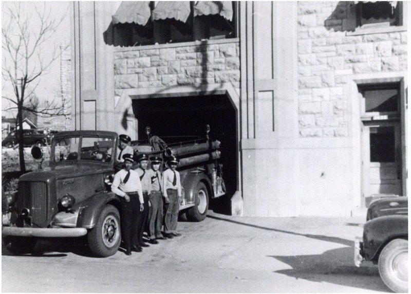 The image shows a group of uniformed men standing next to an open-topped fire truck. The men are wearing light-colored dress shirts, dark pants, dark ties, and dark-colored peaked caps. Four men are standing on the ground to the right of the truck, while a fifth is sitting in the driver's seat. In the background, a large limestone building is visible. The rear of the firetruck is partially backed into an open garage door. To the right of the garage door, there is a smaller closed door. Above the garage, there are three windows, each covered by an awning. On the far right side of the image, the front of a parked car is visible. Past the building on the left side, of the image, trees and some parked cars can be seen further up a hill, with a limestone building partially visible in the background.