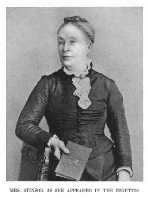 Woman in a dark dress with lace collar, holding a book. Hair tightly wrapped in a printed scarf.