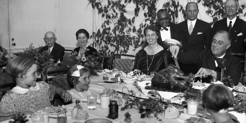 President Franklin D. Roosevelt, accompanied by Eleanor Roosevelt, carves the 1933 Thanksgiving Day turkey with fellow polio patients in the Georgia Hall Dining Room. 