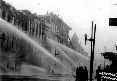 A black and white photo of Stephenson Street in 1928. Firefighters pour streams of water on several buildings as they fight a major fire that affected every building on the block. At lower right are the handwritten words "Hartmann's, Freeport, ILL in white letters.