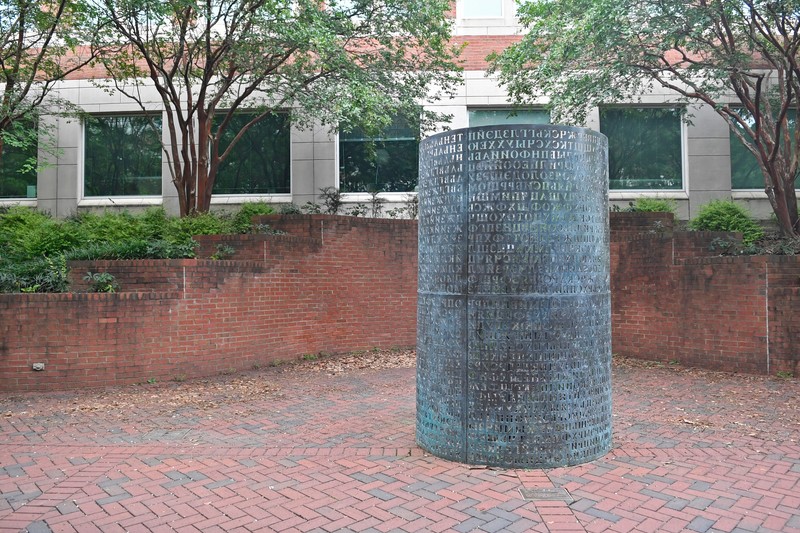 Daytime photo of a bronze, cylindrical sculpture with Cyrillic letters cut out of the surface. Sculpture is installed on a brick patio.