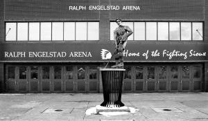 Statue of a hockey player holding his stick at the ready stands on a tall base in front of the Old Ralph.