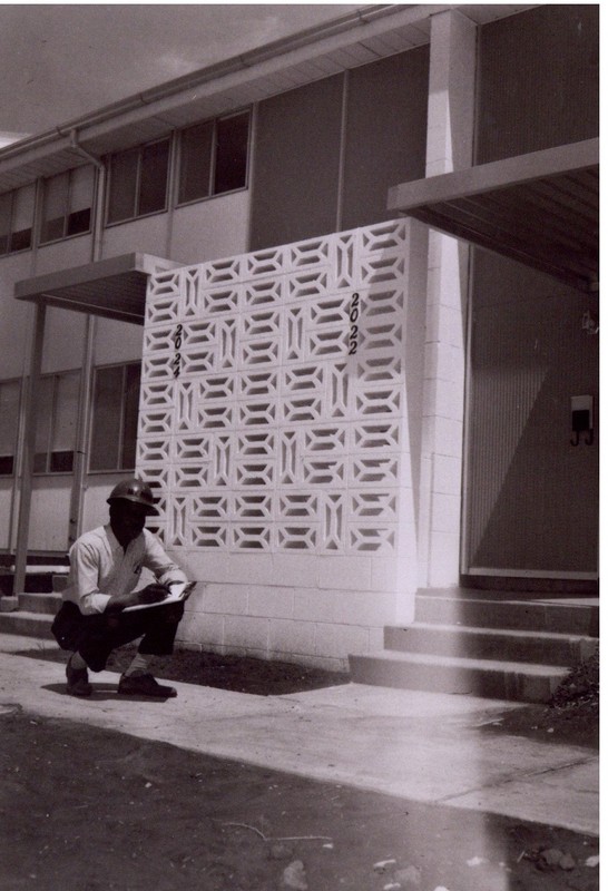  The image shows an African-American man in a light-colored dress shirt, dark pants, dark shoes, and a hardhat. He is holding a clipboard and crouching in front of a decorative cinderblock wall. The wall is part of the porch of two connected townhomes.