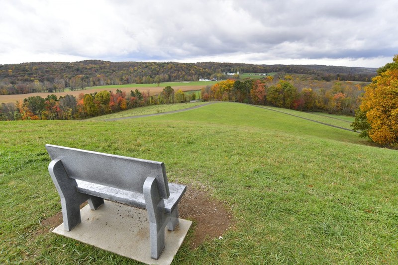 Fall scene with a bench in the foreground overlooking a large valley under a cloudy sky. The white and green buildings of Malabar Farm can be seen far off in the distance.