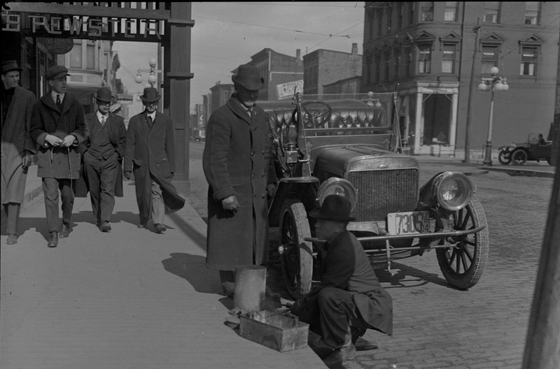 A black and white photo of Stephenson Street circa 1914. At left, a group of four men in winter coats is walking down the sidewalk, passing under a sign for the entrance of The Brewster House hotel. At center a man in a long dark coat is about to get his shoes shined by a man kneeling with a shoeshine kit. Behind them is an early motor car, and another car is passing through the intersection in the background.