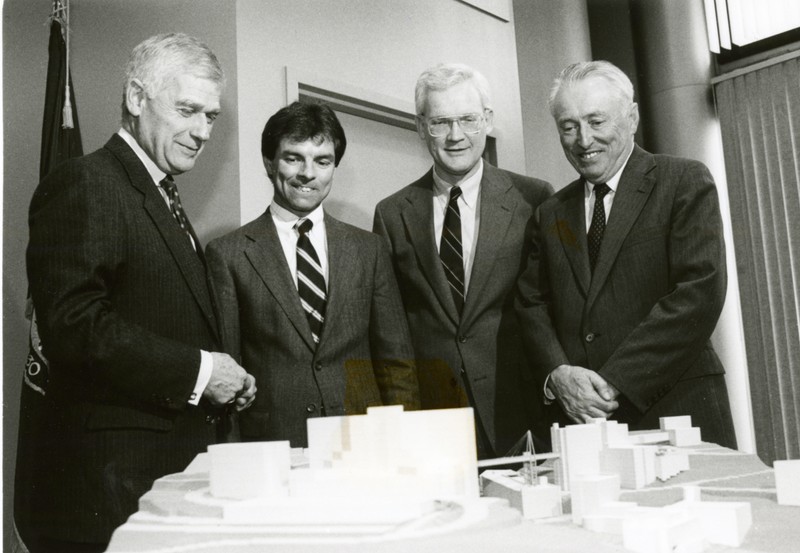Four men in suits smile while looking at an architectural model depicting two hospitals connected by a pedestrian bridge.