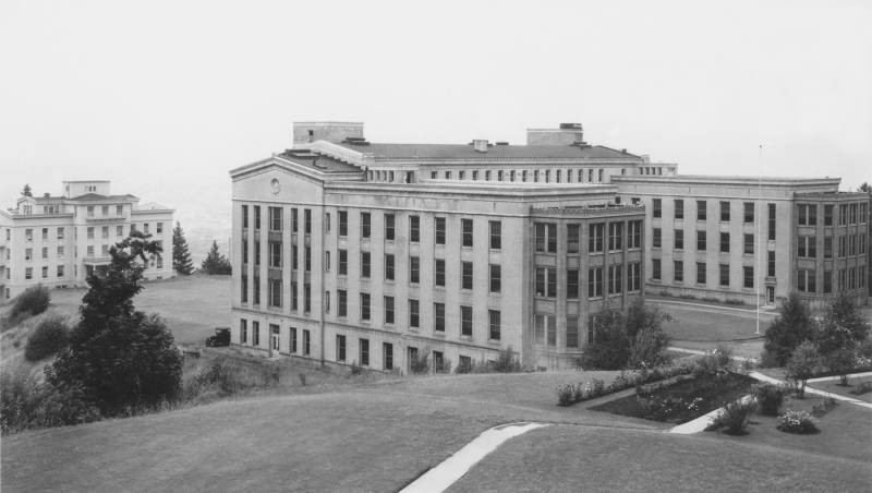 Photograph of (front to back) the Multnomah County Hospital and Emma Jones Hall on the campus of University of Oregon Medical School. The picture has been taken from a position to the southwest of the buildings. The buildings are large, classical style and light colored, and surrounded by a clearing of grass.