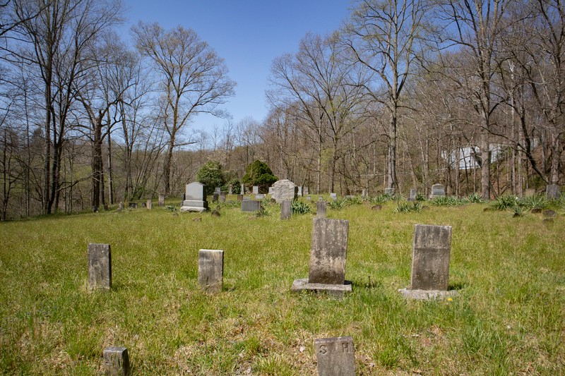 A small cemetery with about 25-30 headstones visible from this vantage point. Most of the headstones are old and weathered. Plants grow up around some of the stones. The sky is brought blue.