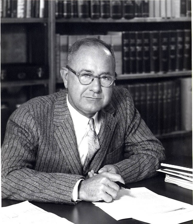 David W. E. Baird sits at a desk wearing a striped suit jacket and tie. He wears small round glasses and has short gray hair. Behind him a bookshelf is visible.
