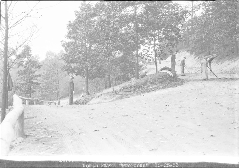 Black and white photo shows a road or trail with a sharp curve. On the lower side there is a man with a fedora and suit looking up at the four men using hand tools on the trail surface. 