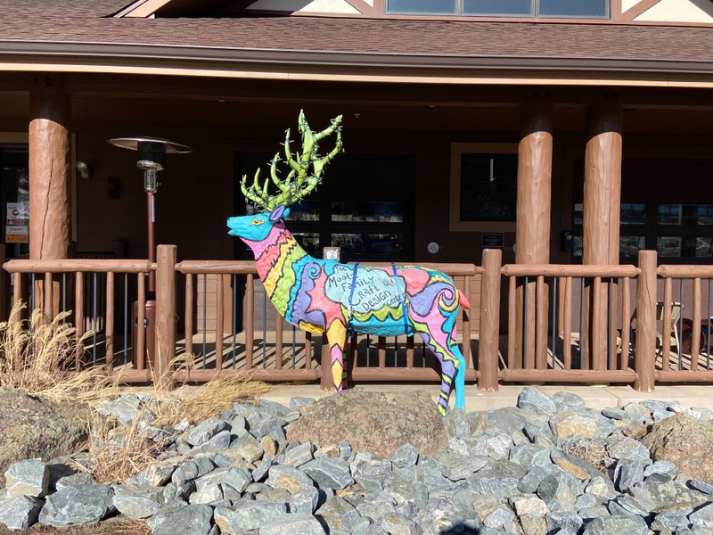 This sculpture of a male elk is painted in various stripes and swirls of bright blue, pink, red, yellow, lime green, and purple. His antlers are lime green and wrapped with lights.