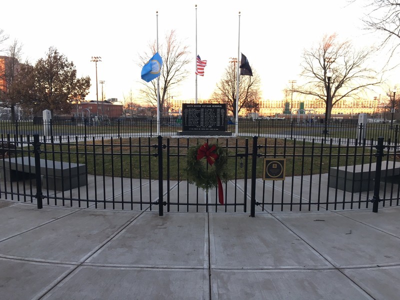 This wide-angle picture of the South Boston Vietnam Memorial shows the Black memorial enclosed within a short gate and framed by the American, POW/MIA, and city of Boston flags. 