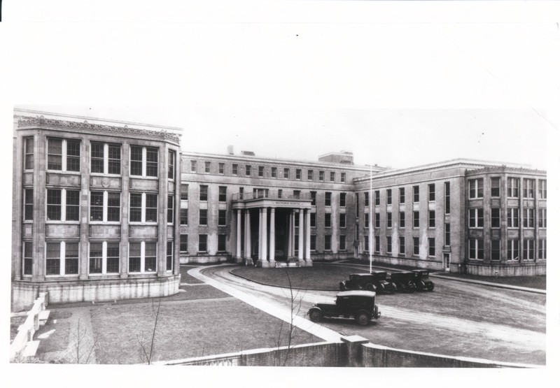 Black and white photograph depicts a large, U-shaped classical style building with large front columns. In front of the building, a driveway circle with several old-fashioned cars is visible.