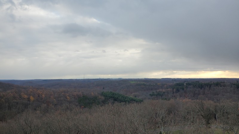 Photo taken from above the tree tops, showing rolling hills and in the far distance the top of a downtown building (US Steel Tower) emerging from the horizon. The tree tops in the foreground are bare, and in the distance some tops of trees are orange or red. In the center is a large stand of evergreen trees. It is a cloudy sky, but the sun is peaking through in the distance on the right.