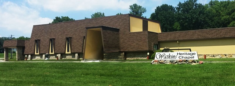 The image shows a view of a large two-story building with shingled siding and cream-colored walls. In the foreground, a large grassy lawn can be seen with a sign on the right side. The sign is white with black text, and is set on a stone brick base. The sign has large cursive text on the right side that reads "WATKINS". On the left side of the sign, black text reads "MEMORIAL CHAPEL" with one word on top of the other. There is a concrete drive between the building and the lawn. In the center of the building, a trapezoid-shaped entryway can be seen. On the left side, tall narrow trapezoid-shaped stained glass windows can be seen on the wall. At the left end of the building, there is an awning supported by two stone pillars covering a small portion of a parking lot. On the right side, there is another single-story wing of the building