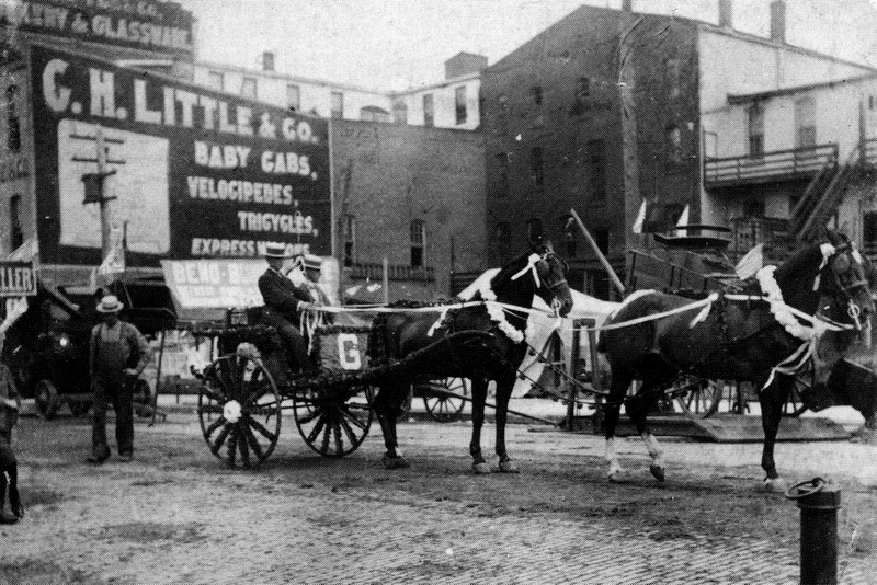 A black and white photo of a street scene at the turn of the 20th Century. Two men with straw hats travel in a carriage decorated for a special event with floral wreaths on the horse's necks and a large G on the front of the carriage. The street is paved with bricks and we see an iron hitching post in the foreground. A man in overalls and a straw hat is crossing the street behind the wagon. On the wall of a building in the right background is an advertisement for C.H. Little & Company's baby cabs, velocipedes and tricycles. Velocipedes were an early type of bicycle.