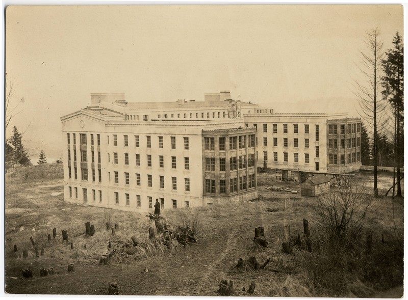 Sepia tone photograph of the Multnomah County Hospital (from the southwest). The building is a large, U-shaped, classical style building. In the foreground, tree stumps and a cleared field are visible.