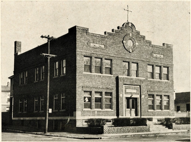 A black-and white photograph of a two-story brick building. The top of the front wall has a Mission-style design with a large circular decoration in the center and a cross mounted to the edge of the roof above it.