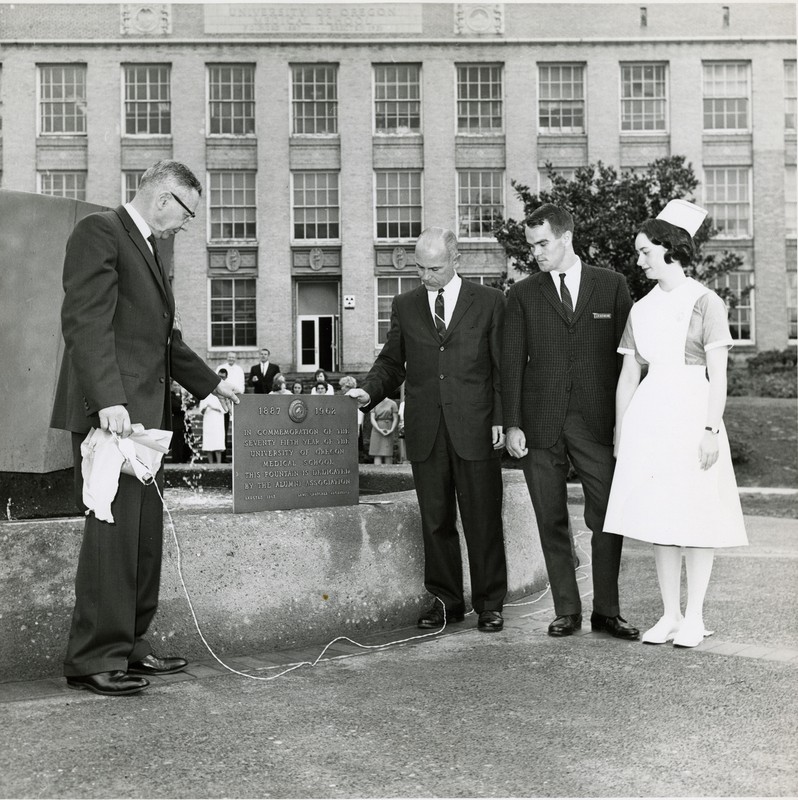 A black and white photo depicts a crowd of people in the background, watching four people standing in front of a fountain looking at a plaque.