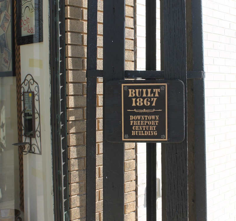 A black iron structure next to a brick wall. On the structure is a bronze plaque reading "Built 1867, Downtown Freeport Century Building"