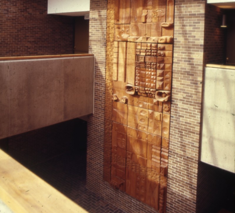 Color negative scan of a two-story modernist wooden sculpture, hanging on a brick wall and visible from a balcony.