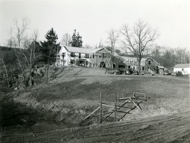 Black and white photo of a large white house surrounded by scaffolding. Four old cars are parked outside and no glass windows are in the house yet.