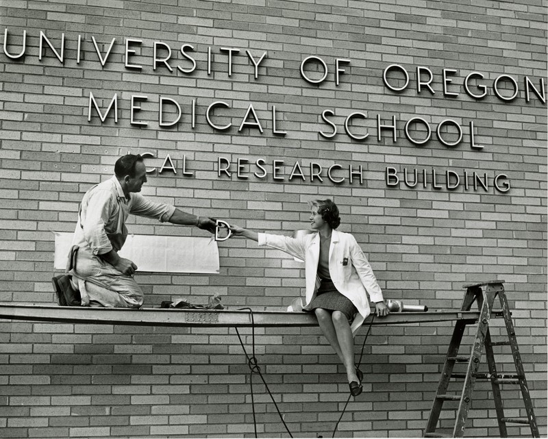 Black and white photograph shows one person handing another the metal letter "D" as they put up the building sign reading "University of Oregon Medical School Medical Research Building"