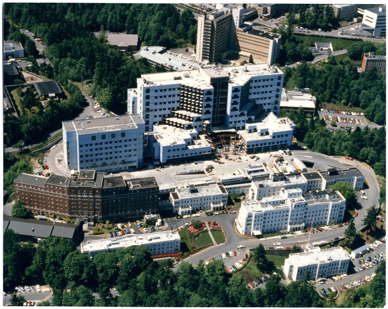 Color photograph of an aerial view of a number of large, 1980s modern hospital buildings under construction against the background of forested canyon and other campus buildings. 