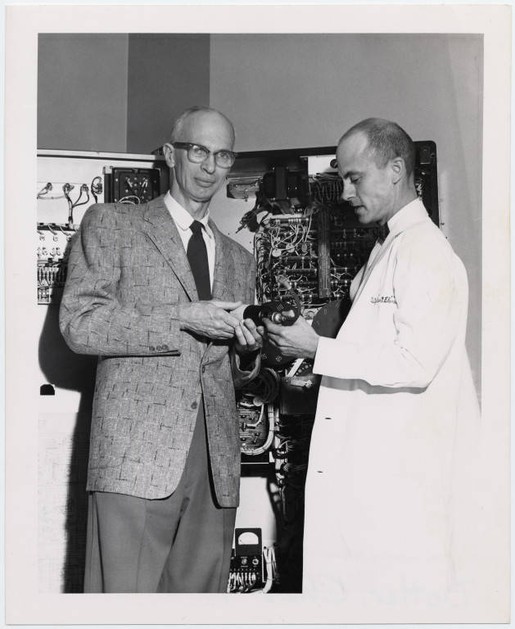 Black and white photograph of M. Lowell Edwards and Charles Dotter in 1956. The two men are standing in front of equipment. 