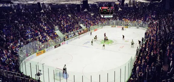 hockey players on ice, with onlookers filling the stadium seating surrounding the rink.