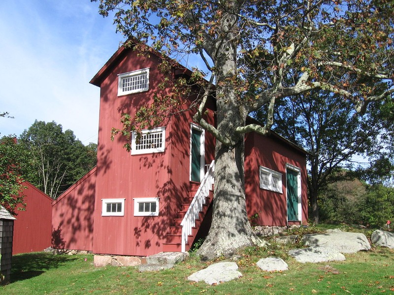 A side shot of Julian Alden Weir's Studio. The three-story building is painted red and has green doors. The windows and outside stair-railings are painted white. Much of the building is obstructed by a large tree. There are stepping stones running through the front yard.